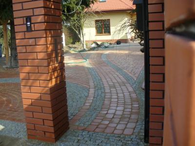 Ułożona z kostki brukowej, zakrzywiona ścieżka prowadząca do domu, widoczna przez ceglany filar bramy wejściowej. Użyto czerwonej i szarej kostki, tworząc dekoracyjny wzór.